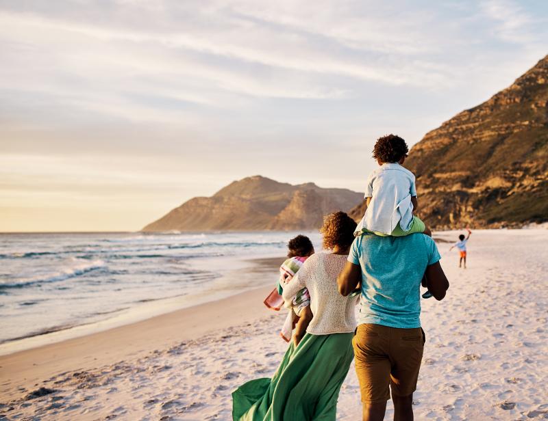 Familie mit drei Kindern läuft den Strand entlang, im Hintergrund das Meer und Felsen