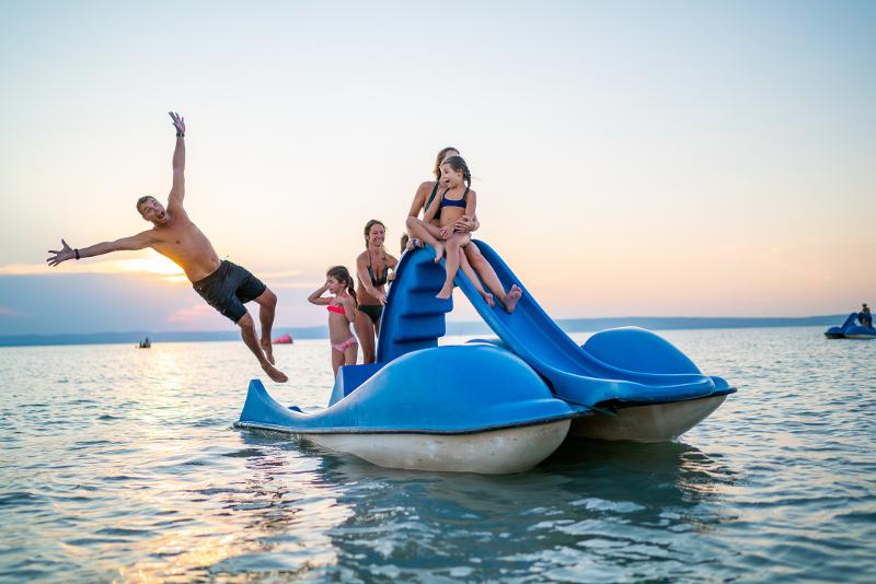 Blaues Tretboot mit Rutsche auf dem Meer im Urlaub für Familien, ein Mann springt ins Wasser und eine Frau mit Kind sitzt auf der Rutsche