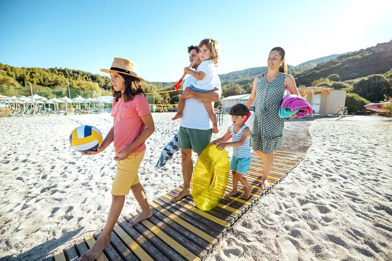 Eine Familie, die im Urlaub mit Kindern Europa Strand auf einem Weg am Strand geht. Sie tragen Strandbekleidung und haben Luftmatratzen, einen Ball und Handtücher im Arm