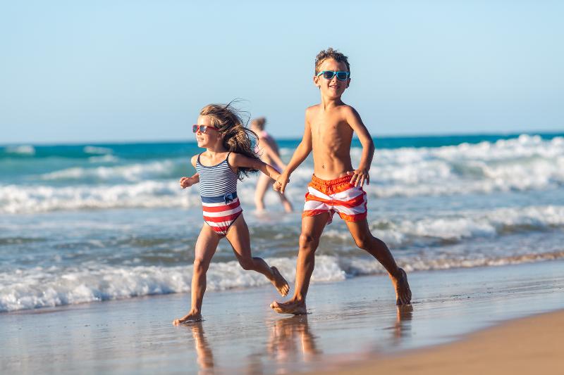 Zwei Kinder, die im Urlaub mit Kindern Europa Strand in Badebekleidung und Sonnenbrillen am Strand entlang rennen, im Hintergrund das Meer mit flachen Wellen