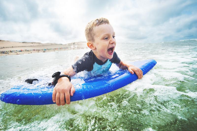 Ein lachender Junge surft auf einem blauen Surfbrett im Frankreich urlaub mit Kindern