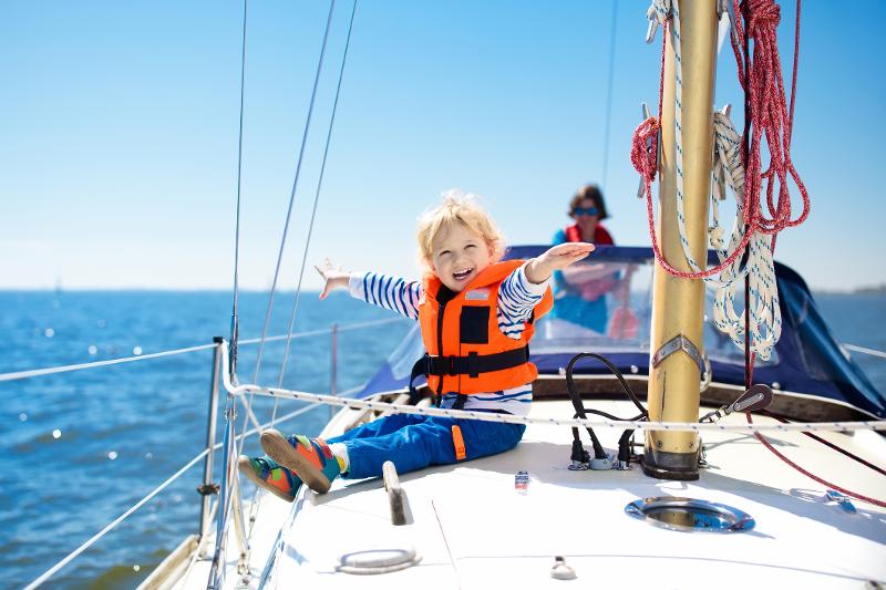 Kind mit orangener Schwimmweste, dass auf einem Boot im Urlaub mit Kindern Holland am Meer sitzt und seine Arme ausstreckt während es lacht