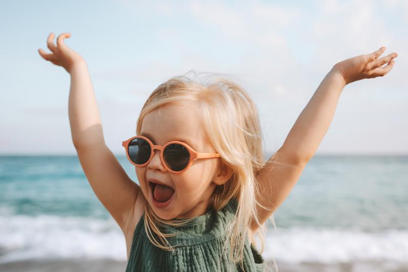 Ein Kind mit Sonnenbrille, das im Urlaub mit Kindern Holland am Meer am Strand ist und sich freut, im Hintergrund das Meer