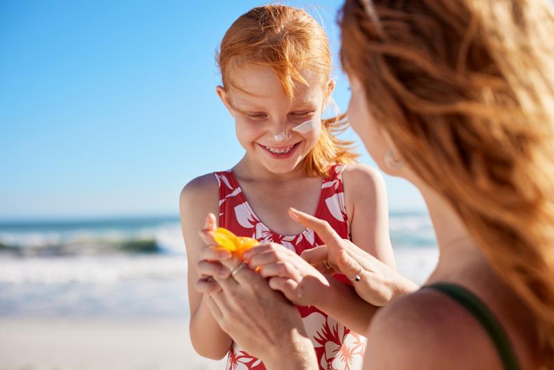 Ein Kind in Badebekleidung, dass im Urlaub mit Kindern Holland am Meer von seiner Mutter Sonnencreme bekommt. Im Hintergrund das Meer