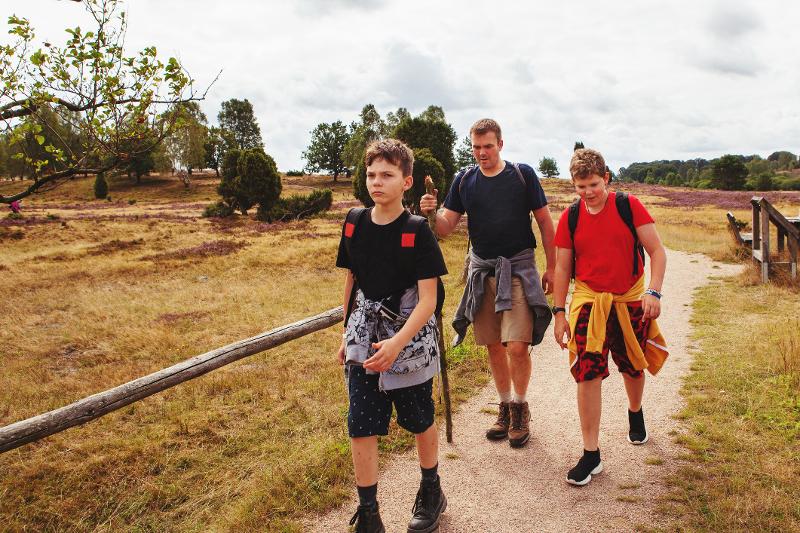 Ein Vater und seine Söhne, die im Urlaub mit Kindern Niedersachsen einen Weg entlang gehen. Um sie herum sieht man die Lüneburger Heide mit Wiesen und Bäumen