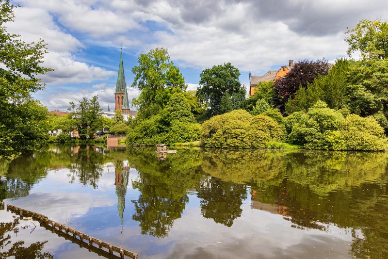 Ein Blick auf Oldenburg in einem Stadtpark mit großem Teich, Bäumen und einer Kirche im Urlaub mit Kindern Niedersachsen