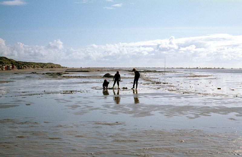 Eine Familie, die im Urlaub mit Kindern Niedersachsen im Watt stehen und buddeln. Im Hintergrund sieht man die weite des Strandes und eine Pfützen