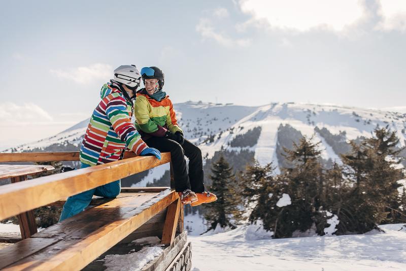 Zwei Personen in bunter Skikleidung sitzen lachend auf einer Holzbank mit Blick auf die verschneiten Pisten.
