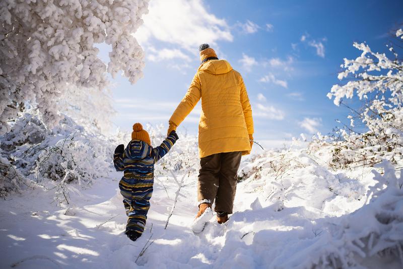 Mutter und Kind spazieren Hand in Hand durch eine verschneite Landschaft unter einem strahlend blauen Himmel.