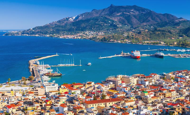Aussicht auf die Hafenstadt mit Häusern, dem Meer mit Booten und einem Berg im Zakynthos Urlaub mit Kindern
