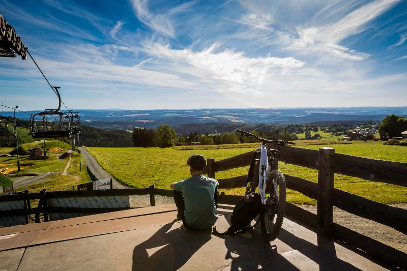 Der Himmel ist strahlend blau und die Berglandschaft des Vogtlandes saftig grün. Ein Radfahrer genießt die Aussicht.