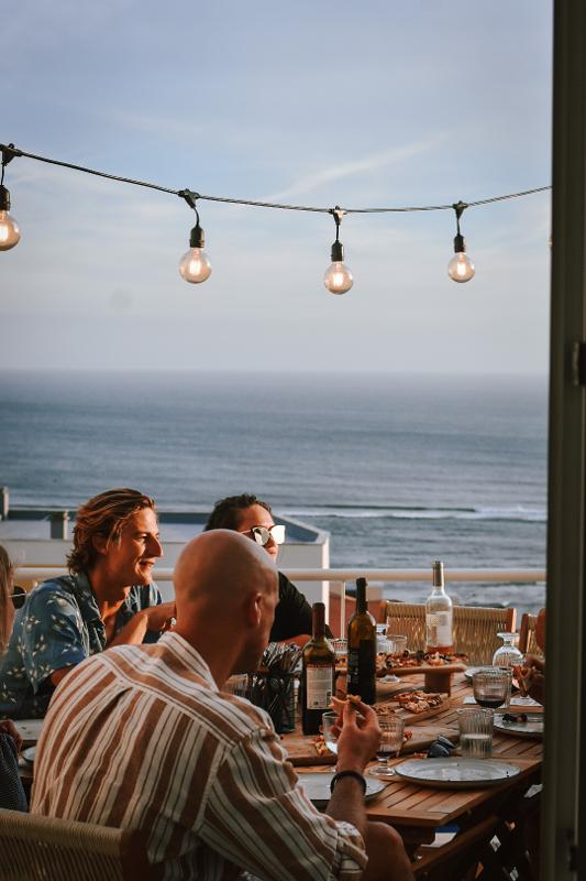 Menschengruppe am Esstisch auf einer Terrasse mit Meerblick im Familienurlaub Surfen