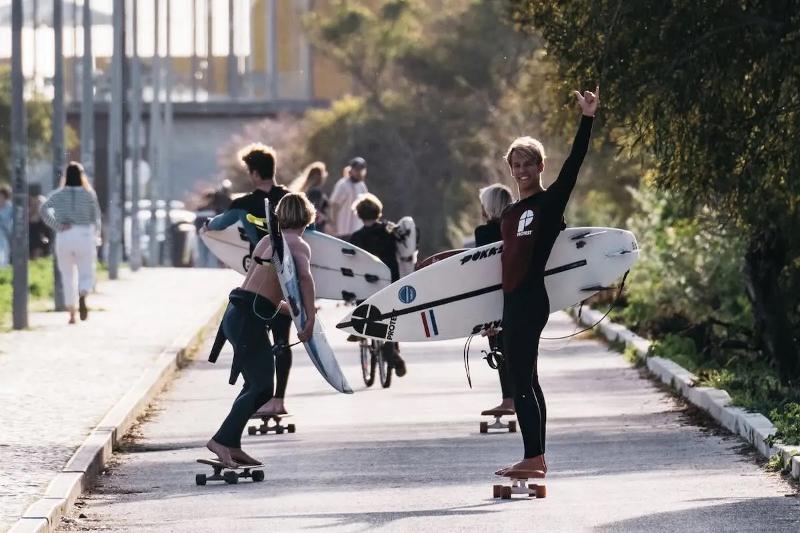 Gruppe auf Skateboards mit Surfbrettern unterm Arm im Familienurlaub Portugal