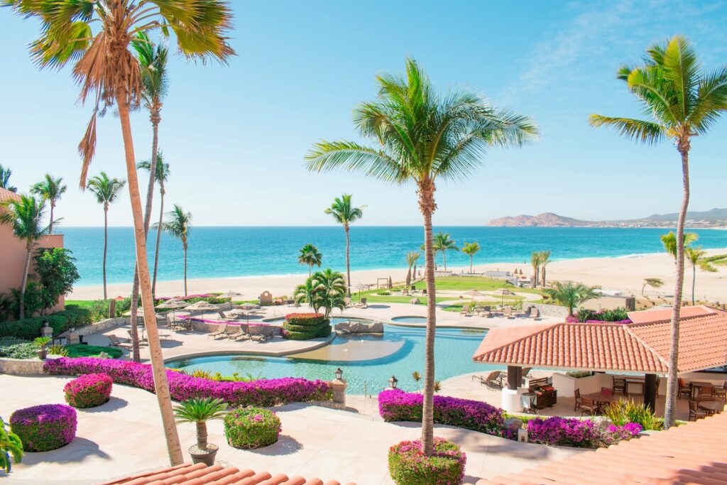 Große Poolanlage mit Palmen, pinken Blumen, Gebäuden am Strand und dem blauen weiten Meer im Hintergrund bei schönem Wetter im Urlaub mit erwachsenen Kindern