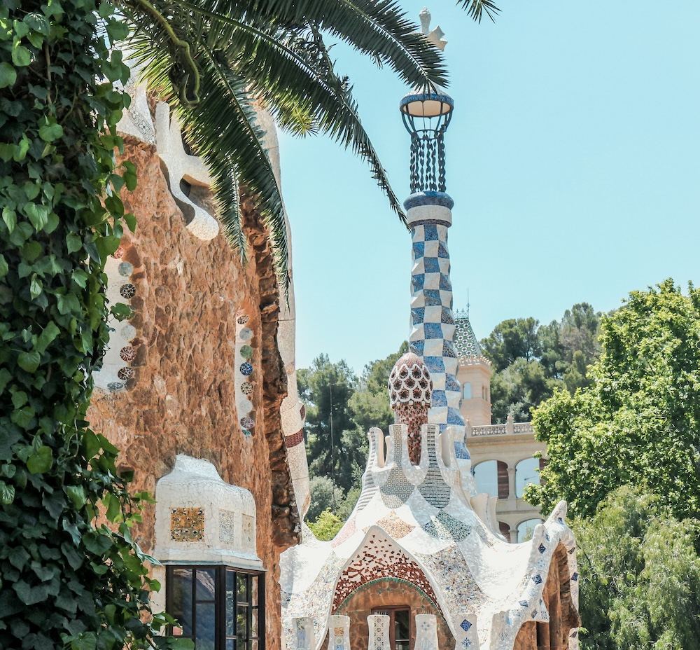 Der Park Güell in Barcelona mit grüner Palme im Vordergrund und einem für den Park typischen Haus mit Säule im blau weiß design bei schönen Wetter im Urlaub mit erwachsenen Kindern