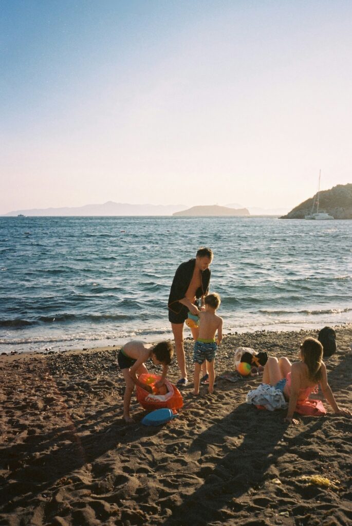  Eine Familie die am Strand spielt, im Hintergrund sieht man das Meer und noch einen Teil einer Klippe im Sonnenschein