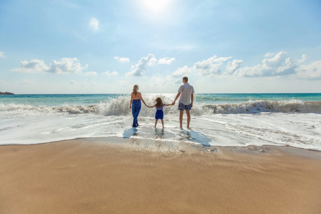 Wohin Urlaub mit Kindern? Eine Familie, die sich am Strand an den Händen hält und die dem Meer zugewandt ist. Es tauchen vermehrt Wellen auf und die Sonne scheint.