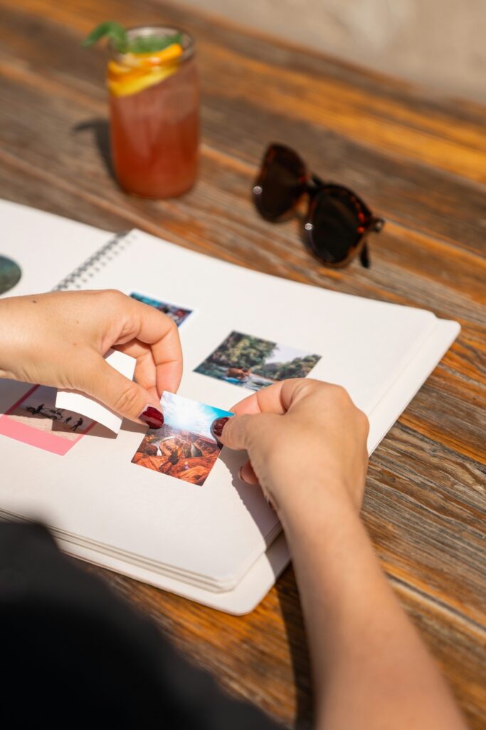 Tipps Familienurlaub: Fotoalben. Ein Mensch klebt Fotos in ein Buch. Außerdem sieht man eine Sonnenbrille und ein Getränk.