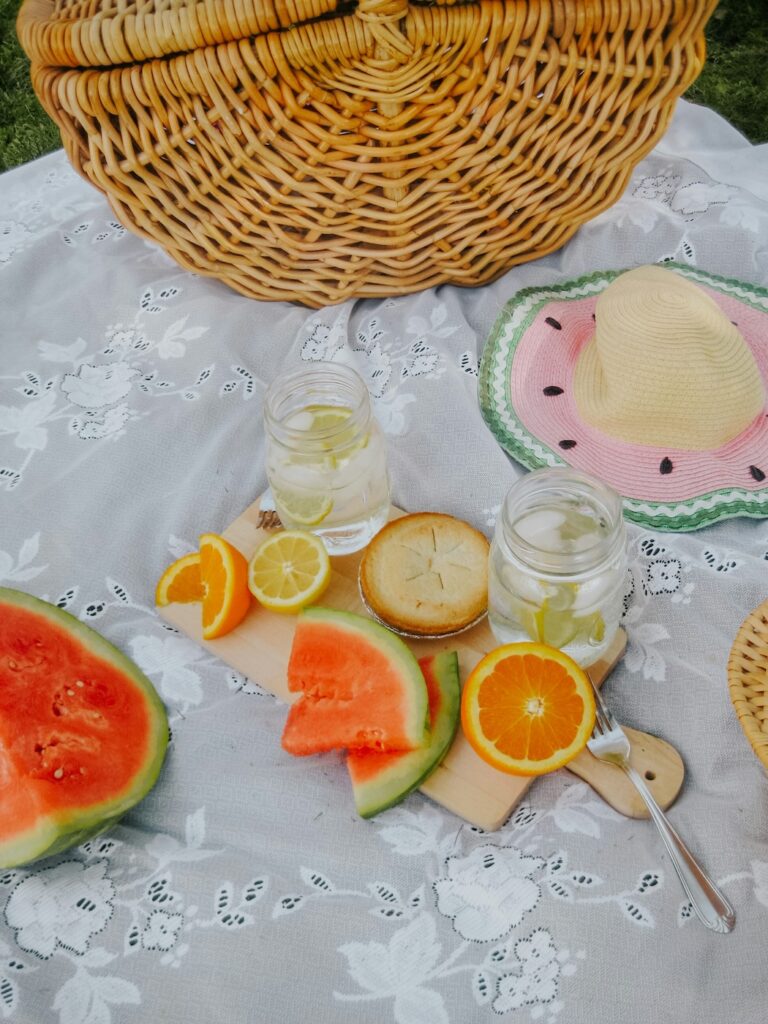 Tipps Familienurlaub: eine weiße Picknickdecke und ein Korb. AUf einer Platte sind Wassermelonenstücke, Kuchen und Getränke. Außerdem sieht man einen Hut und Besteck.