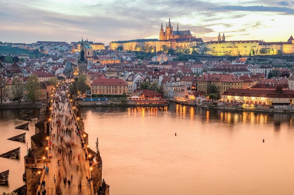 Urlaub mit erwachsenen Kindern - wohin? Ein Überlick auf Prag mit einer Brücke, dem Fluss und der Stadt. Im Hintergrund kann man auf einem Berg das Schloss in der Dämmerung sehen.