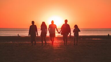 Alles rund um Tipps Familienurlaub. Eine Familie am Strand. Im Hintergrund sieht man das Meer und den Sonnenuntergang.